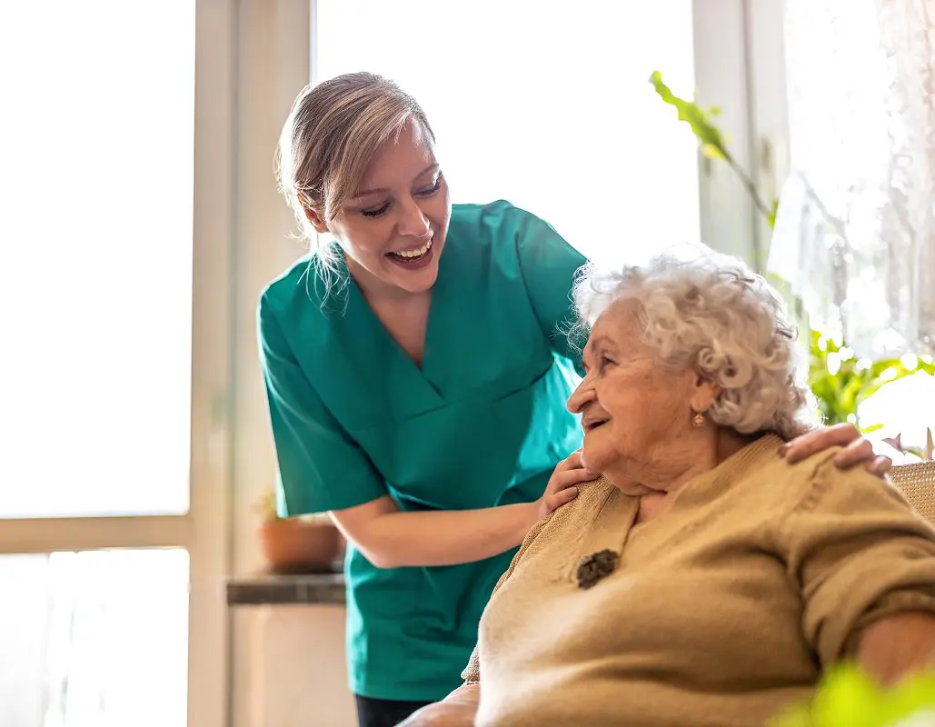 A caregiver in teal scrubs smiling and gently holding the shoulder of an elderly woman with curly white hair, who is sitting and smiling back; they are in a bright room with large windows and green plants.