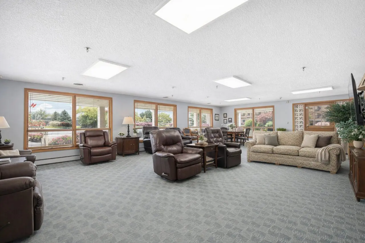 A spacious lounge area with large windows, several brown leather recliners, a beige patterned sofa, side tables with lamps, and a dining table with chairs. The room is well-lit with ceiling lights and has a view of trees outside.