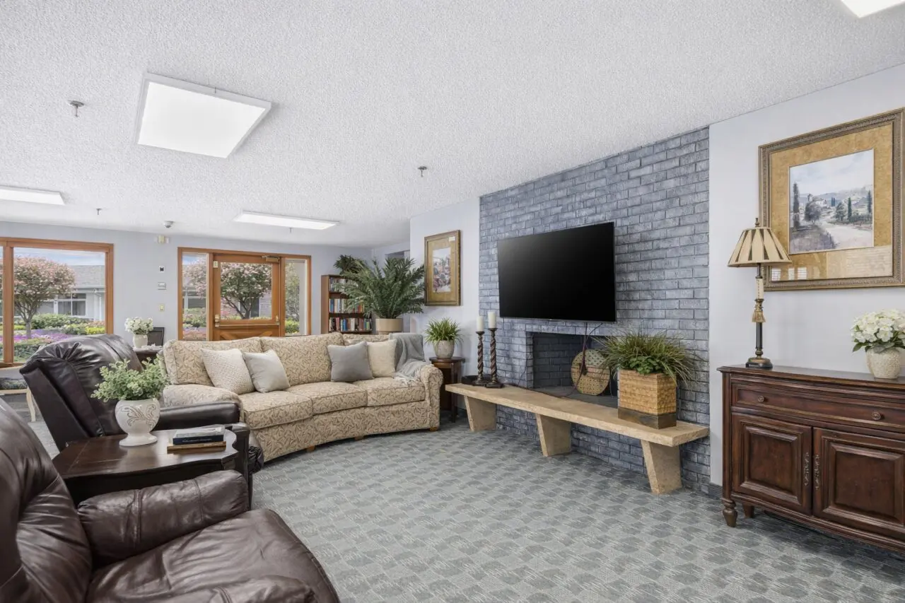 A cozy living room with a beige patterned sofa and dark brown leather chairs, a large flat-screen TV mounted on a gray brick wall, framed artwork, a wooden sideboard with a lamp and flower vase, and large windows showing trees and a building outside.