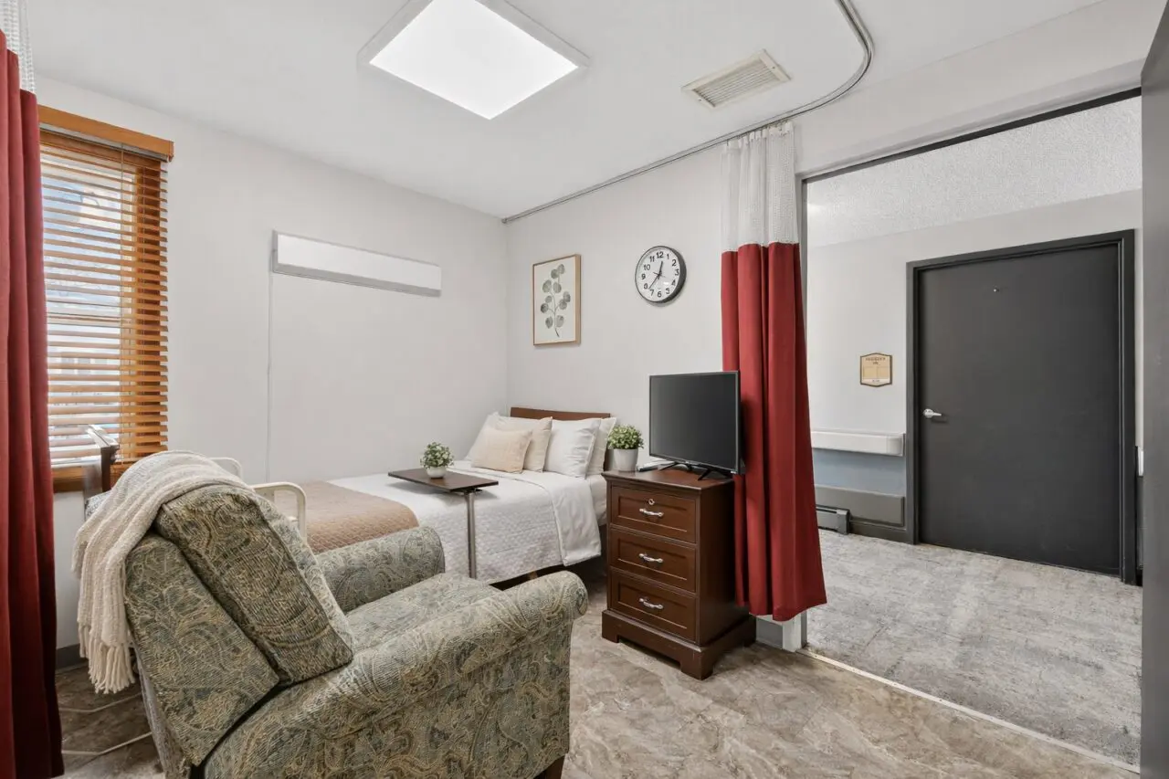 A cozy room with a bed and a small wooden dresser holding a TV, a patterned armchair with a blanket, a window with wooden blinds and red curtains, and wall decorations including a clock and framed artwork.