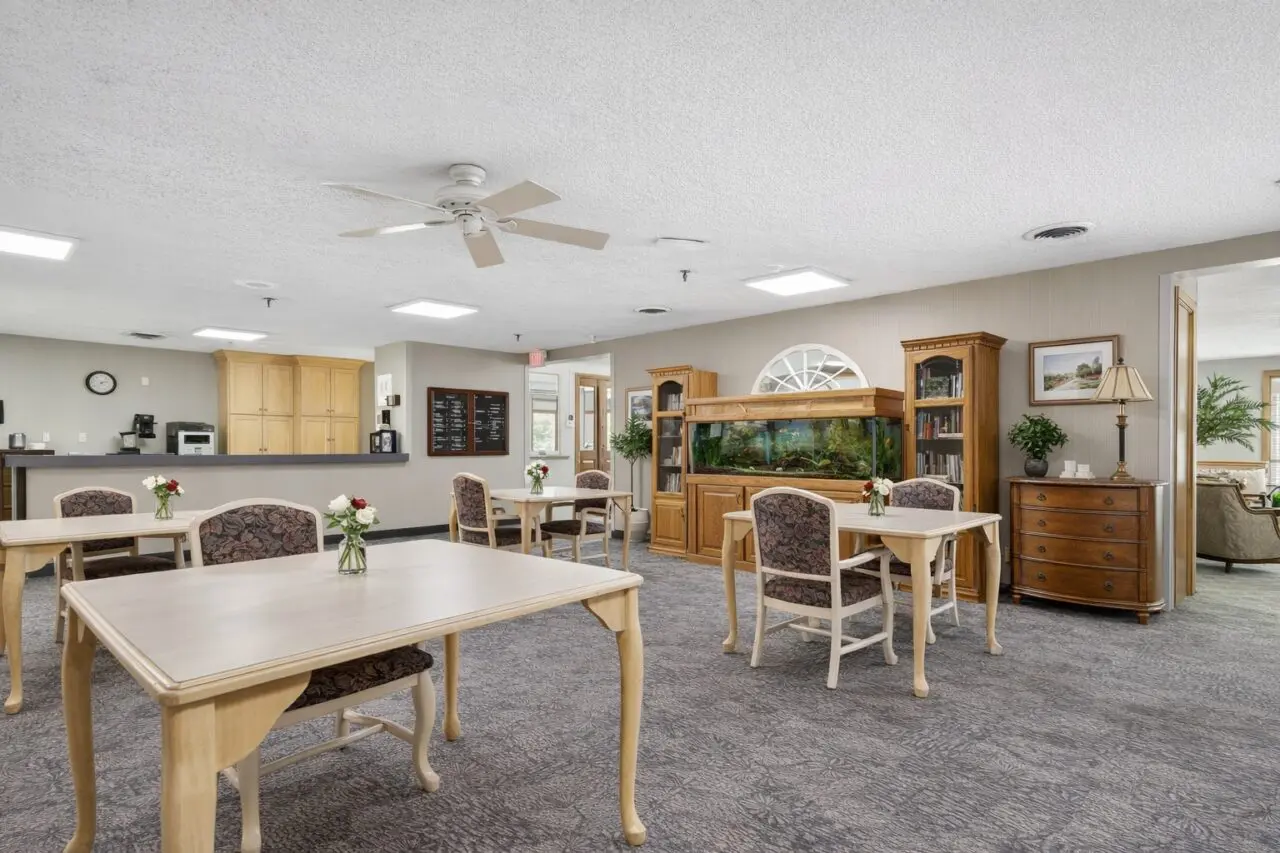 A spacious common area with light-colored wooden tables and chairs, decorated with small flower arrangements, and a large fish tank against the wall. In the background, there is a kitchenette with cabinets, a clock, and a chalkboard menu. The room has a ceiling fan and carpeted floor.