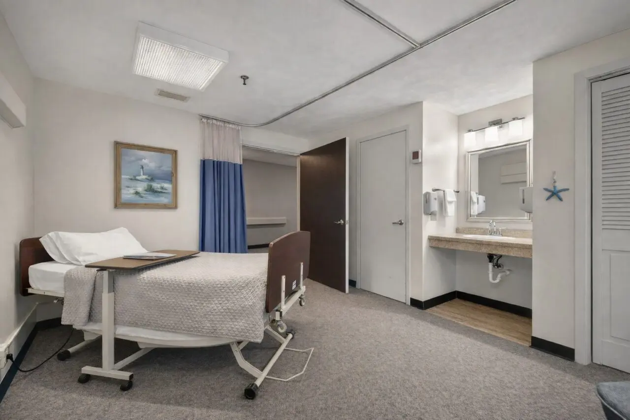 A hospital room with a bed on wheels, white bedding, and a pillow, positioned against a white wall with a framed painting of a lighthouse and ocean. There's a blue and white curtain partially covering an area behind the bed. To the right, there is a sink with a mirror, paper towel dispensers, and a starfish decoration on the wall. The room has gray carpeted flooring and a ceiling with fluorescent lighting.