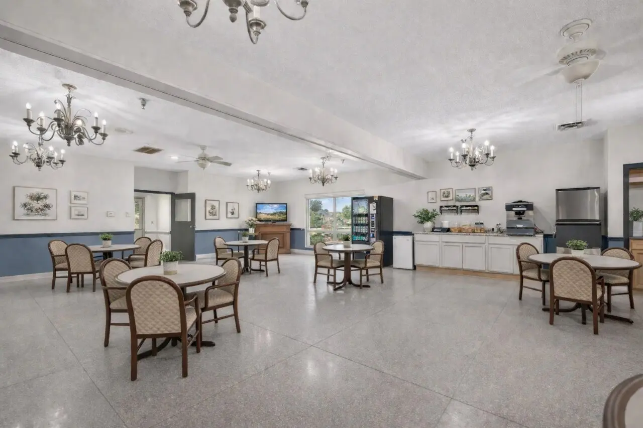 A spacious dining area with several round tables surrounded by cushioned chairs, chandeliers hanging from the ceiling, framed artwork on the walls, a vending machine, a TV, and a refreshment station with a coffee machine and supplies.