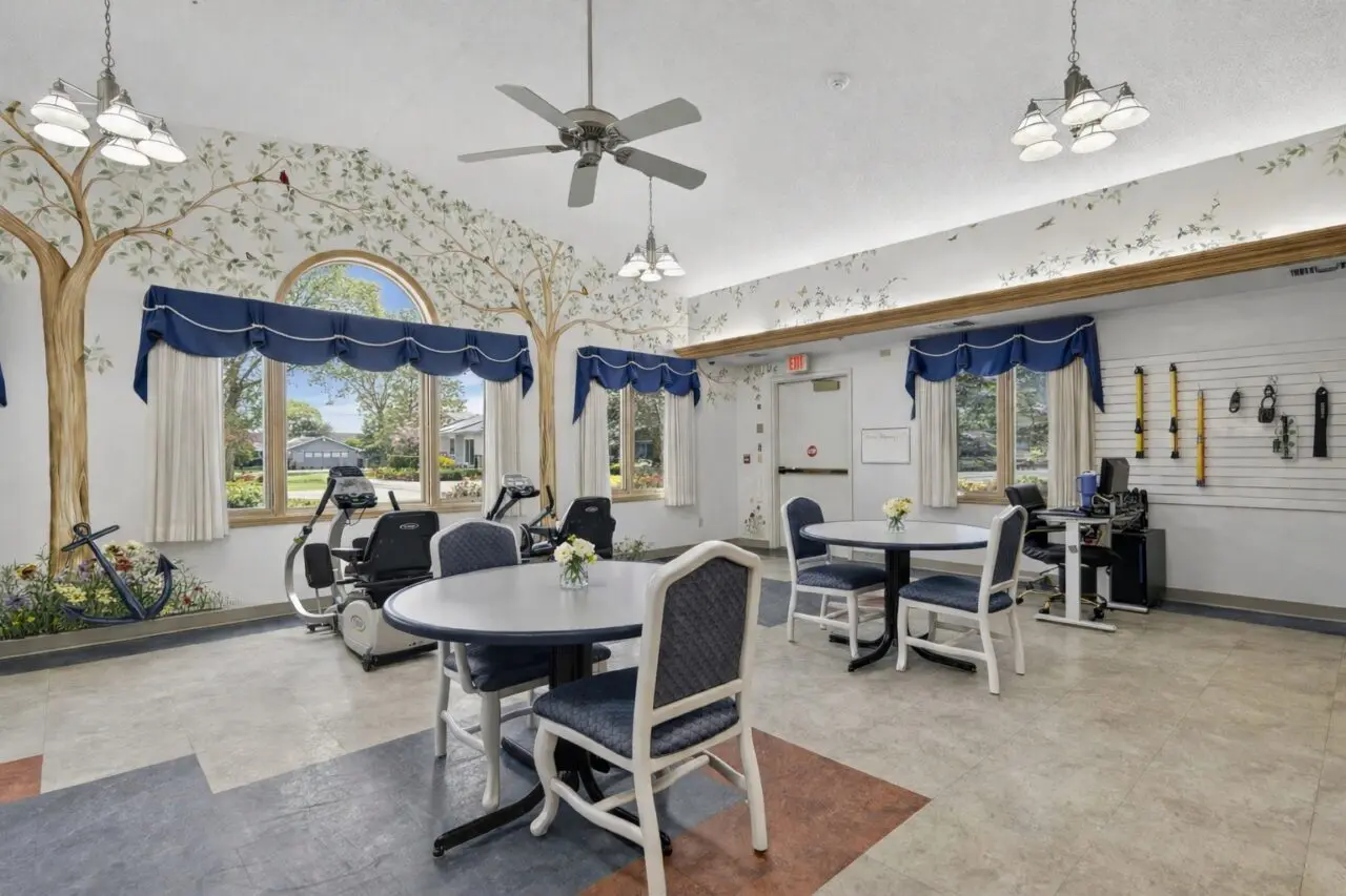 A bright activity room with large windows, blue valances, and white curtains, decorated with tree and leaf patterns on the walls. There are round tables with chairs, exercise equipment near the windows, and a wall with medical and therapy tools. A ceiling fan and multiple light fixtures hang from the ceiling.