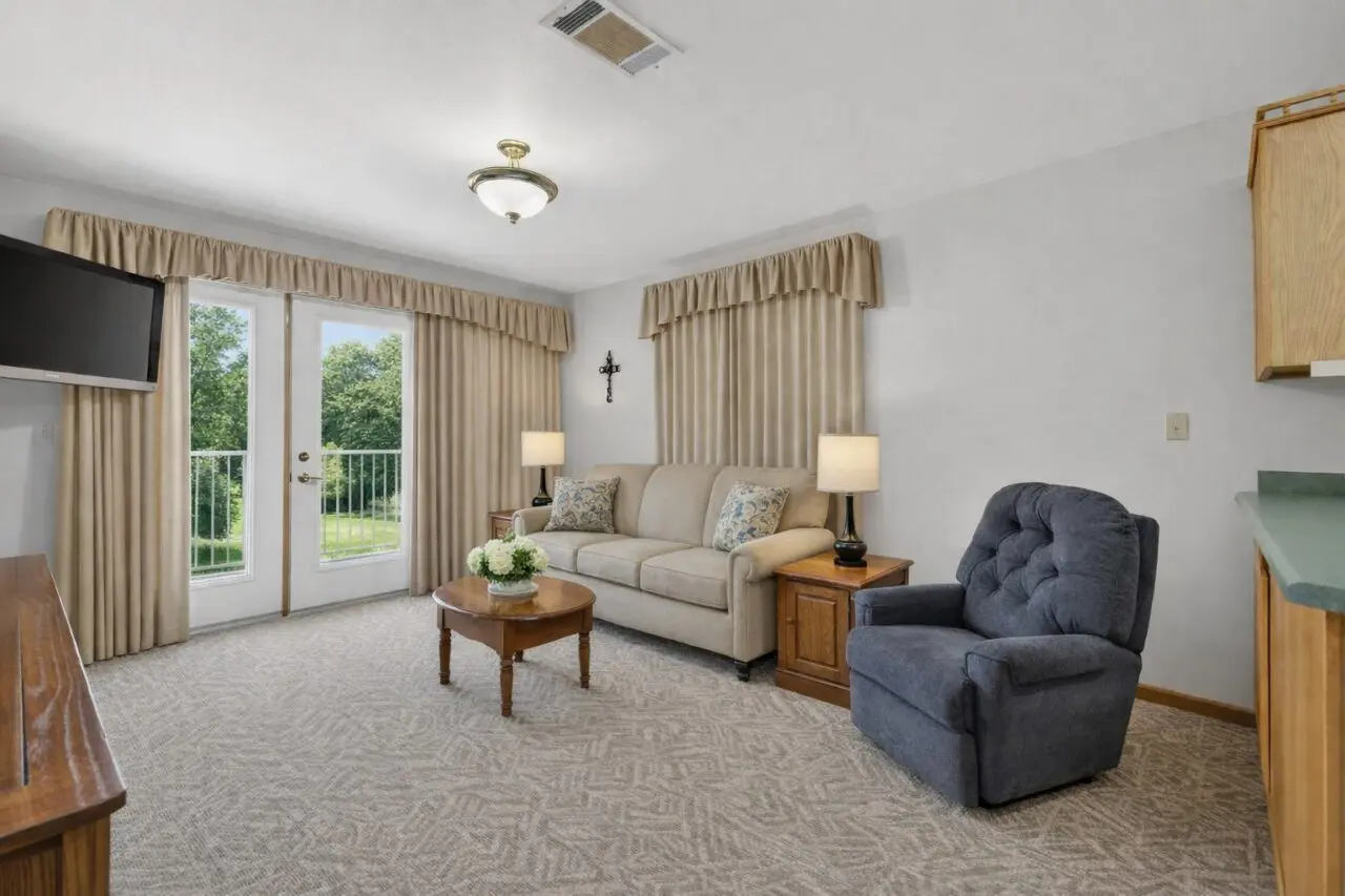 A cozy living room with a beige sofa, two side tables with lamps, a blue armchair, a small round wooden coffee table with a flower arrangement, and a wall-mounted TV. There are large glass doors with beige curtains leading to a green outdoor view. A cross hangs on the wall above the sofa. The room has a neutral color scheme and carpeted floor.