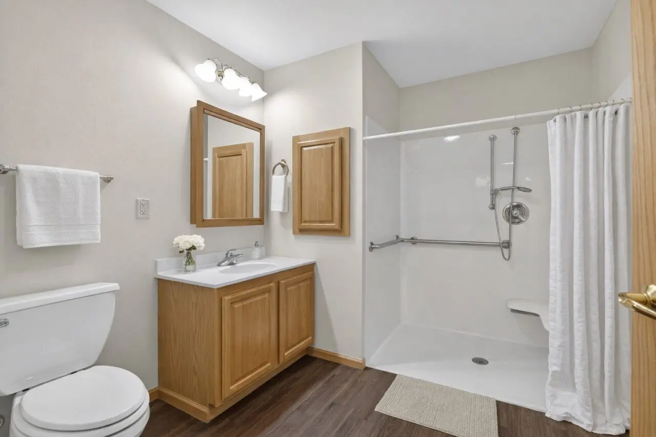 A bathroom with a white toilet, a wooden vanity with a white countertop and a vase of white flowers, a wooden medicine cabinet, a mirror, a towel rack with a white towel, and a walk-in shower with a white curtain, a grab bar, and a handheld showerhead.