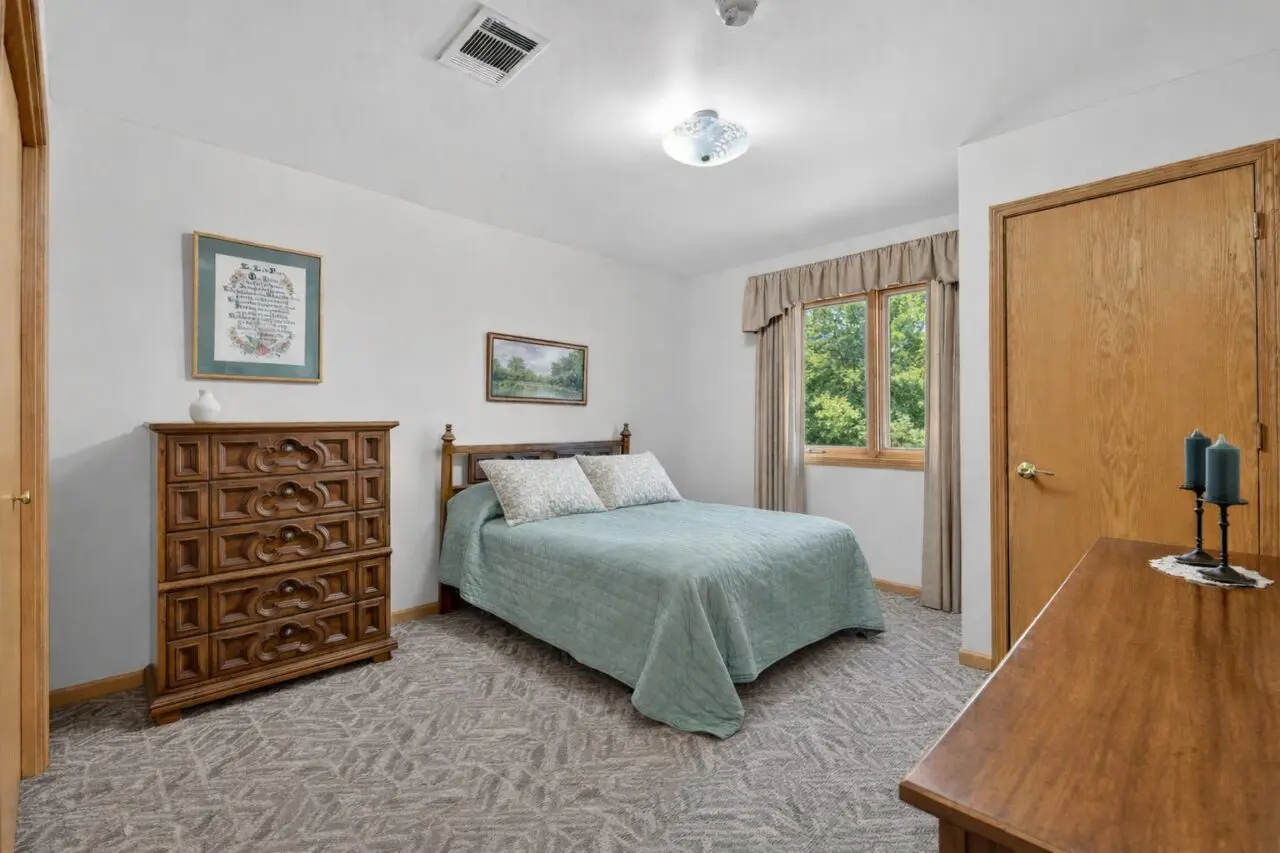 A bedroom with a bed covered in a light green quilt, two matching pillows, a wooden headboard, and a window with beige curtains showing green trees outside. There is a wooden dresser with a small decorative item on top, and a wooden door on the right side. The ceiling has a light fixture and a vent.