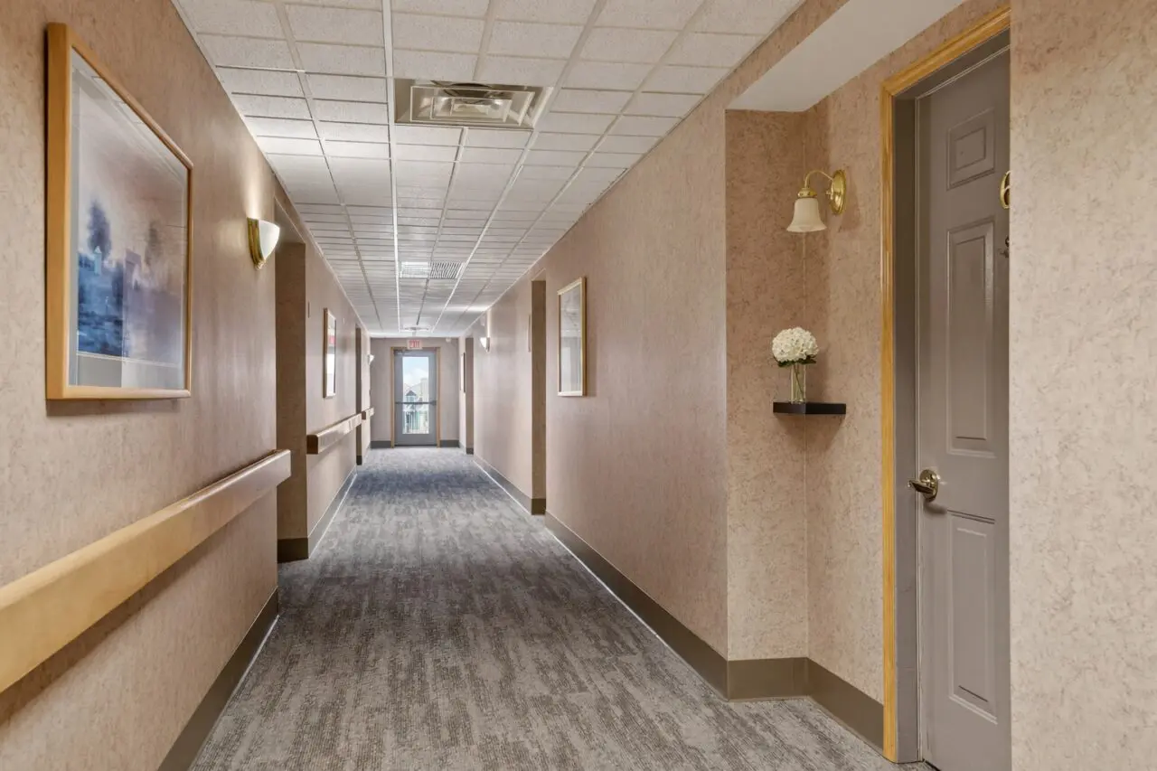 A long, well-lit hallway in a care center with beige patterned walls, framed artwork, wall-mounted light fixtures, and a gray door with a gold handle on the right. The hallway has a gray carpet with a subtle pattern and a ceiling with white acoustic tiles and air vents. At the end of the hallway, a glass door with a balcony view is visible.
