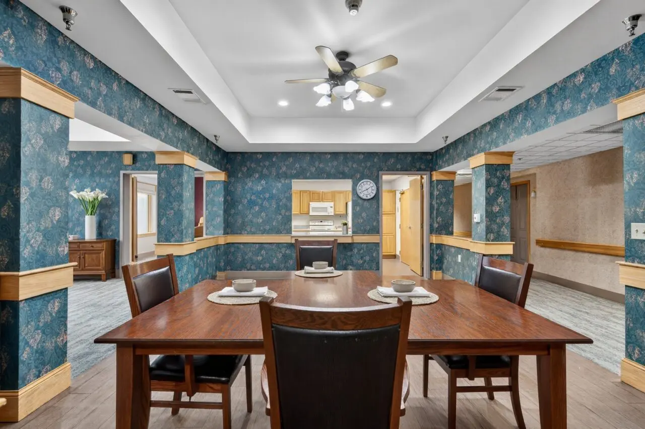 A dining area with a wooden table set for four, with bowls and placemats, in front of a blue floral wallpapered wall with gold trim. There is a ceiling fan above, and a kitchen opening in the background.