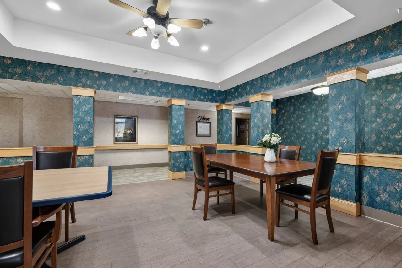 A dining area with a wooden table and four chairs, a vase with white flowers on the table, blue patterned wallpaper, wooden trim, and a ceiling fan with lights.
