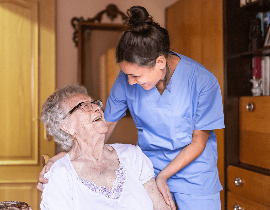 A nurse in blue scrubs is smiling and leaning over a seated elderly woman with gray hair and glasses, who is also smiling and looking up at the nurse. They are sharing a joyful moment in a room with wooden furniture.